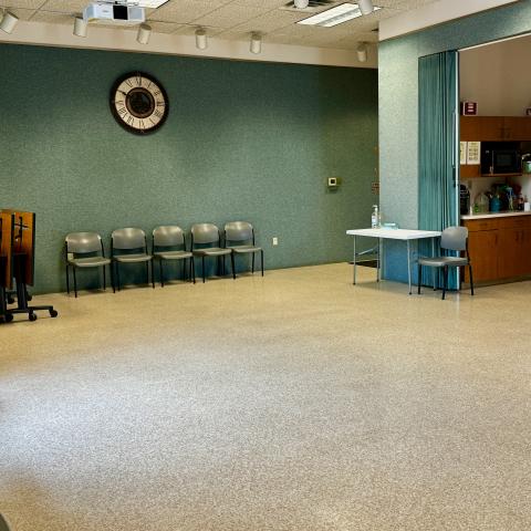 Large room with tables store to the left, chairs along wall, kitchenette to the right.  The walls are green and there is a clock on the wall.